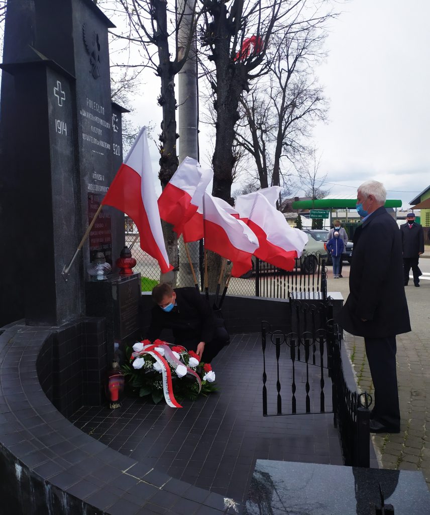 Dwaj mężczyźni przed pomnikiem. Jeden z nich składa kwiaty. W tle biało-czerwone flagi.