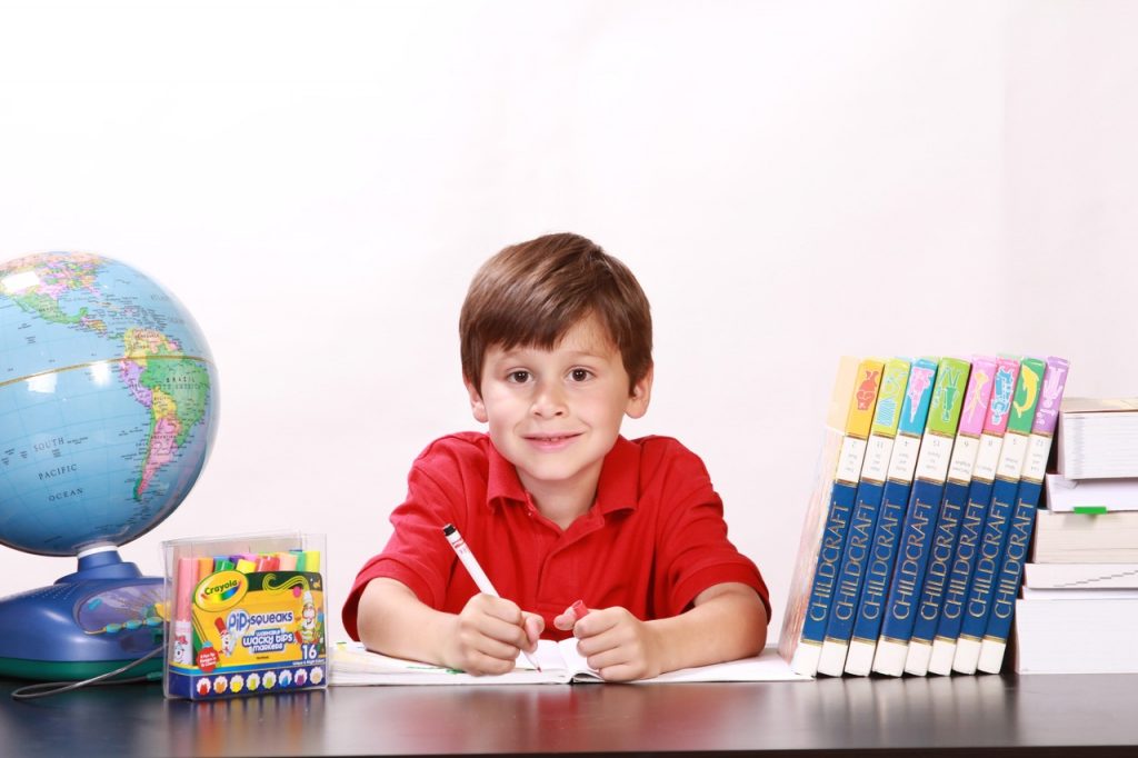 Chłopiec ubrany w czerwoną koszulę siedzący z długopisem w ręku przy biurku. Na biurku leżą przybory szkolne takie jak globus, kredki i książki.