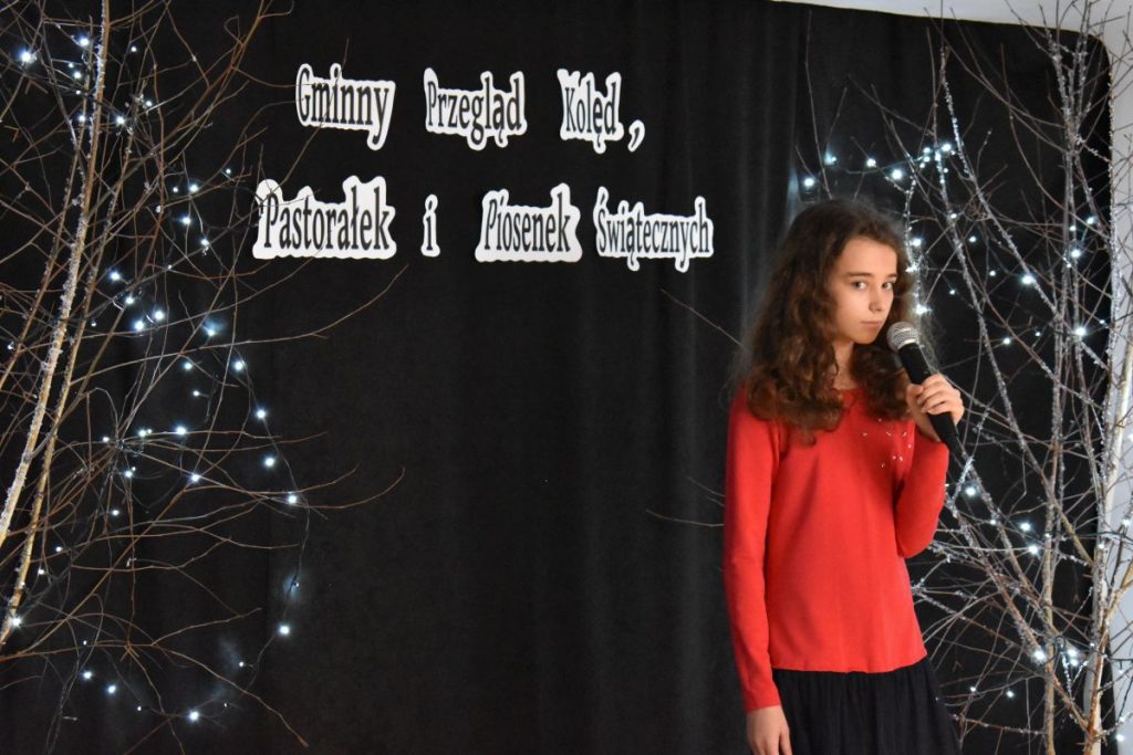 10. Dziewczyna w czerwonej bluzce i czarnej spódnicy trzyma mikrofon w ręku na tle czarna kotara na niej napis Gminny Przegląd Kolęd, Pastorałek i Piosenek Świątecznych i drzewka ze światełkami