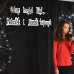 10. Dziewczyna w czerwonej bluzce i czarnej spódnicy trzyma mikrofon w ręku na tle czarna kotara na niej napis Gminny Przegląd Kolęd, Pastorałek i Piosenek Świątecznych i drzewka ze światełkami