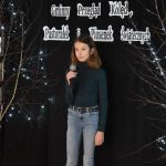 11. Na tle czarnej kotary dziewczynka w granatowym golfie i jasnych spodniach trzyma mikrofon w ręku obok niej stoją drzewka ze światełkami.