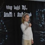 12. Dziewczynka o blond włosach z białą opaską, w białym golfie i beżowych spodniach trzyma mikrofon w ręku. W tle czarna kotara drzewka ze światełkami