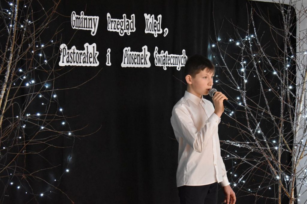 14. Chłopiec w białej koszuli i czarnych spodniach śpiewa do mikrofonu który trzyma w ręku za nim czarna kotara, drzewka ze światełkami