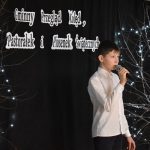 14. Chłopiec w białej koszuli i czarnych spodniach śpiewa do mikrofonu który trzyma w ręku za nim czarna kotara, drzewka ze światełkami