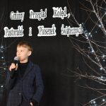 17. Chłopiec w marynarce w kratę trzyma mikrofon w reku na czarnym tle obok gałązki z lampkami