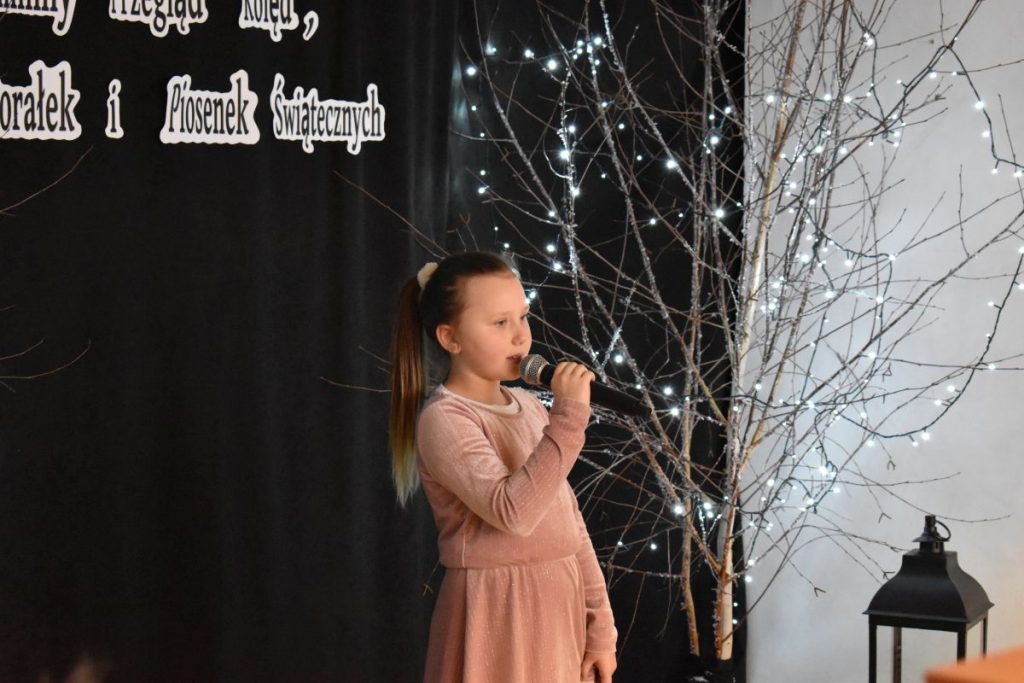 19. Dziewczynka w jasnej sukience śpiewa do mikrofonu na czarnym tle drzewko ozdobione lampkami