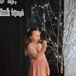 19. Dziewczynka w jasnej sukience śpiewa do mikrofonu na czarnym tle drzewko ozdobione lampkami