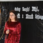 Kobieta w czerwonej sukience i okularach stoi trzymając mikrofon w ręku. W tle czarna kotara i regał z książkami po obu stronach drzewka ze światełkami