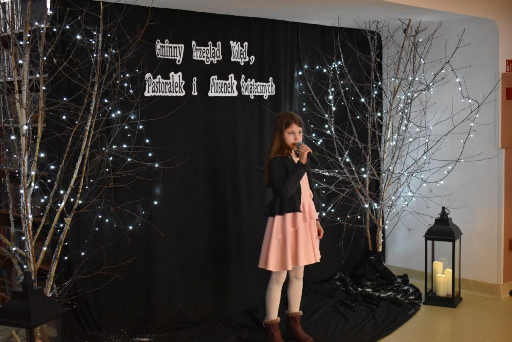 21. Dziewczynka w jasnej sukience i czarnym sweterku trzyma w ręku mikrofon po obu stronach stoję brzozy ozdobione lampkami w tle czarna kotara na podłodze stoi świecący się lampion