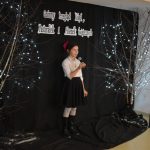 23. Na czarnym tle jest napis Gminny Przegląd Kolęd, Pastorałek i Piosenek Świątecznych, pomiędzy dwoma drzewkami ze światełkami stoi dziewczynka z czerwoną kokardą we włosach w białej bluzce, czarnej spódnicy i czarnych kozakach. W reku trzyma mikrofon, na podłodze stoją dwa lampiony.