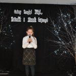 24. Dziewczynka w białej bluzce i spódnicy w kratkę trzyma w rekach mikrofon. W tle czarna kotara dwa drzewka ozdobione światełkami na podłodze lampion