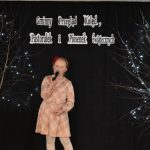 25. Pomiędzy dwoma drzewkami ozdobionymi światełkami stoi dziewczynka w jasnej sukience w kratę na głowie ma czerwoną kokardę. W tle czarna zasłona na niej napis Gminny Przegląd Kolęd, Pastorałek i Piosenek Świątecznych, oraz regał z książkami.