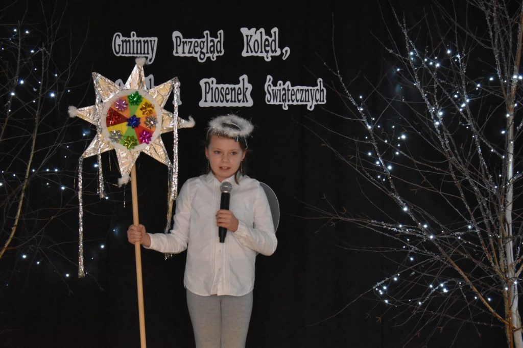26. Dziewczynka w białej bluzce i szarych spodniach na głowie ma aureolę, w jednym ręku trzyma mikrofon a w drugim gwiazdę kolorową na długim kiju. Na czarnym tle gałązki ozdobione światełkami