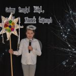 26. Dziewczynka w białej bluzce i szarych spodniach na głowie ma aureolę, w jednym ręku trzyma mikrofon a w drugim gwiazdę kolorową na długim kiju. Na czarnym tle gałązki ozdobione światełkami