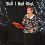 3. Na czarnym tle widoczny napis Gminny Przegląd Kolęd, Pastorałek i Piosenek Świątecznych po obu stronach gałązki na których wiszą światełka. Pomiędzy nimi stoi kobieta przed nią pulpit z pomarańczową teczką. W ręku trzyma mikrofon, ubrana jest w bluzkę w kwiaty z białym kołnierzykiem i czarną spódnicę.