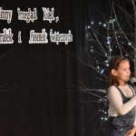 32. Na tle czarnej kotary i drzewek ozdobionych światełkami, dziewczynka w ogrodniczkach i białej bluzce śpiewa do mikrofonu.