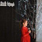 34. Dziewczyna z kucykiem w czerwonej sukience z mikrofonem w reku, w tle drzewko ozdobione światełkami i czarna kotara