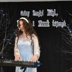 4. Dziewczyna w niebieskiej sukience z białą opaską na głowie grająca na keyboardzie, w tle czarna kotara i napis Gminny Przegląd Kolęd, Pastorałek i Piosenek Świątecznych i drzewka ze światełkami