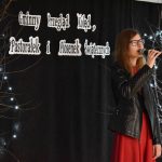 7. Młoda kobieta w okularach, czerwonej sukience i czarnej marynarce trzyma mikrofon w ręku. W tle czarna kotara po obu stronach drzewka ze światełkami.