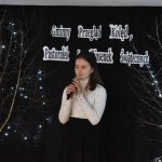 9. Na tle czarnej kotary stoi dziewczyna w białej bluzce i czarnej spódnicy trzyma oburącz mikrofon po obu stronach stoją drzewka ze światełkami.