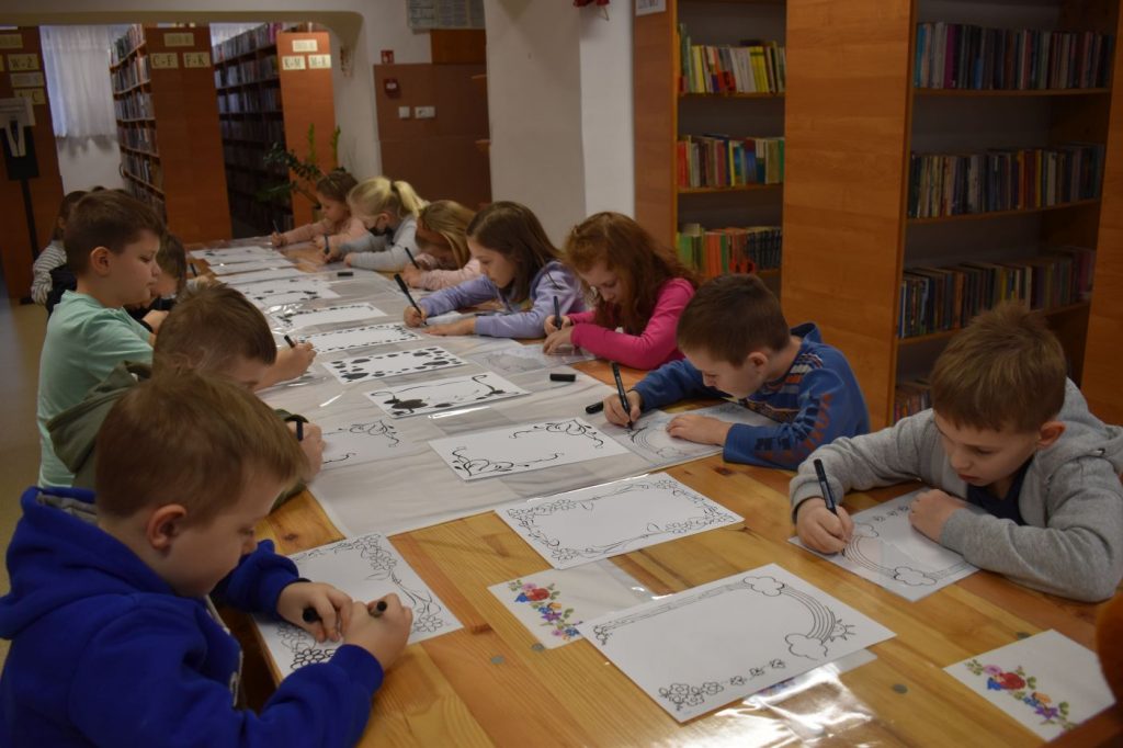 Przy długim stole dzieci rysują na kartkach, w tle regały z książkami