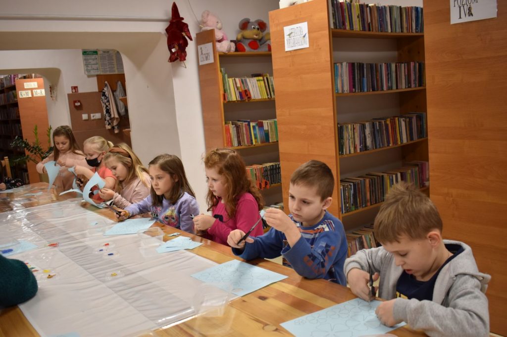 Dzieci siedzą przy stole wycinają kwiatki z kolorowego papieru, za nimi półki z książkami