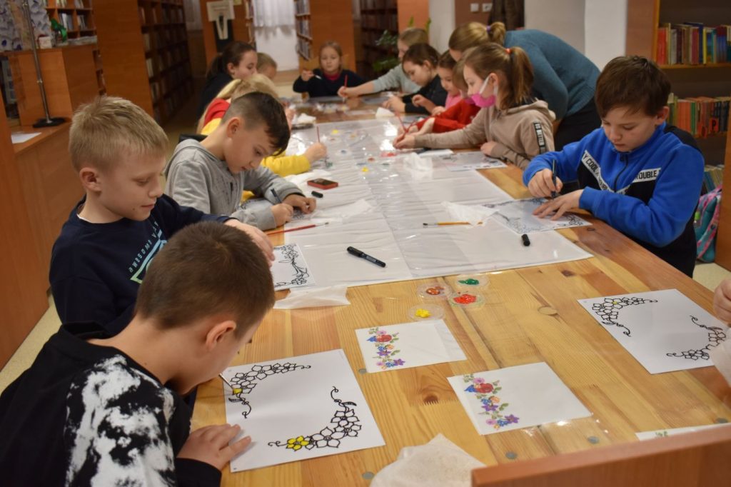 Grupka dzieci maluje farbami kwiatki przy długim drewnianym stole w oddali widać biblioteczne regały z książkami