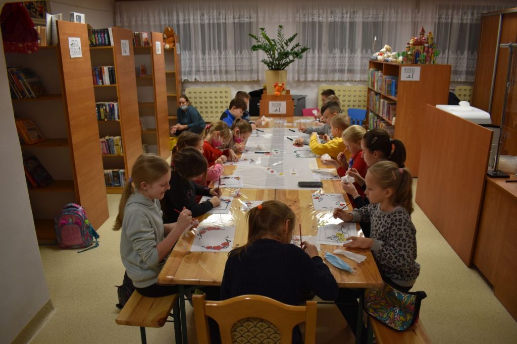W pomieszczeniach biblioteki stoi długi stół przy którym siedzą chłopcy i dziewczęta malując kolorowe rysunki. W tle regały z książkami, na środku stoi kwiatek a za nim widać okna