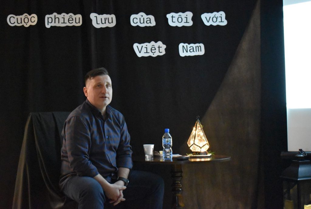 Mężczyzna siedzący przy stoliku na którym stoi lampka w kształcie stożka i butelka wody, w tle czarna kotara na którym jest napis w języku wietnamskim