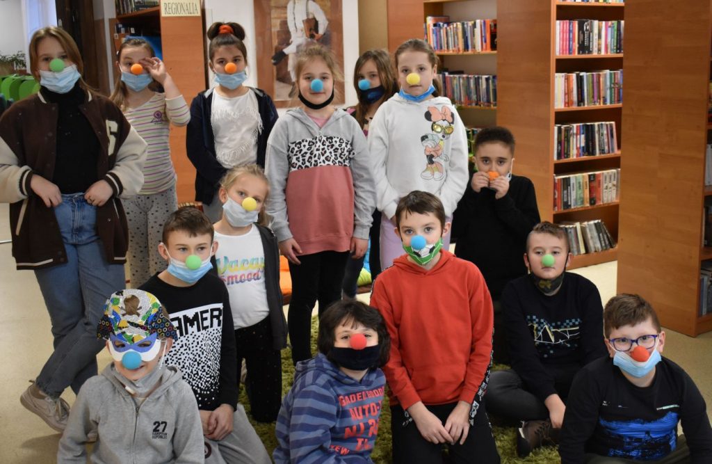 W bibliotece grupka dzieci z kolorowymi noskami za nimi regały z książkami