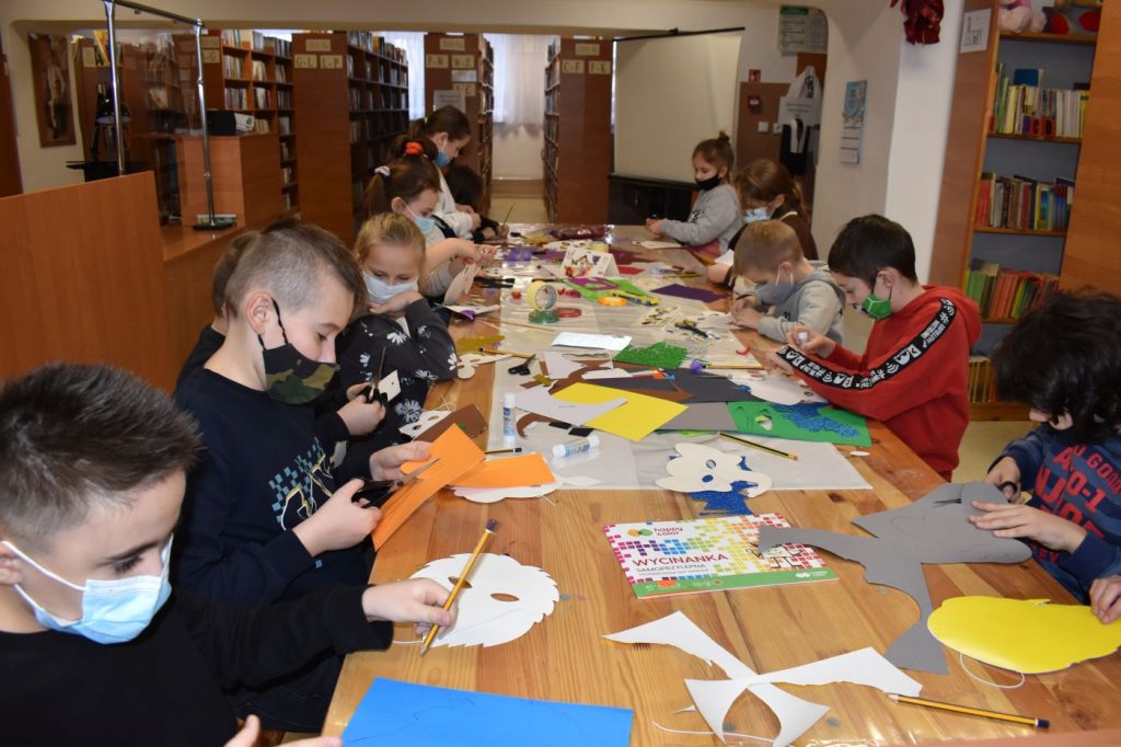 W bibliotece przy długim stole siedzą dzieci i wycinają z kolorowych papierów ozdoby na maski karnawałowe. W tle regały z książkami.