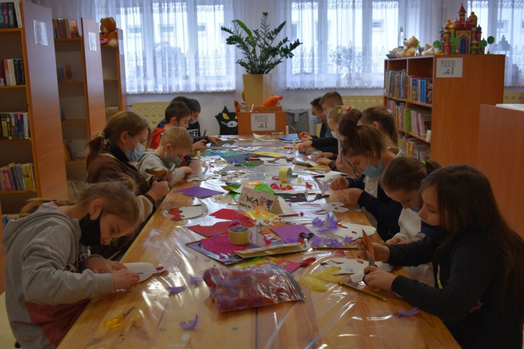 Przy długim stole siedzą dzieci wycinają kolorowe ozdoby z papieru. W tle regały z książkami