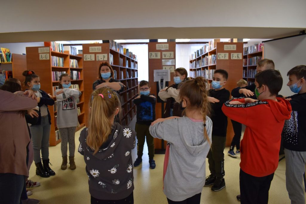 Stojące w kółku dzieci trzymają ręce oparte na łokciach za nimi regały z książkami