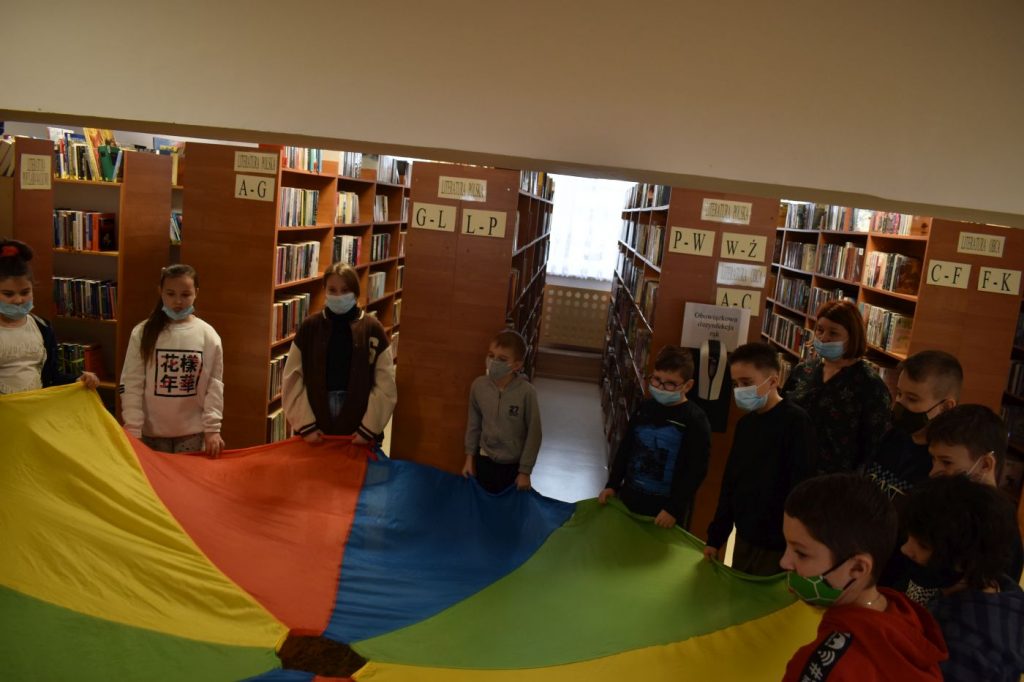 Kolorowa chusta którą trzymają dzieci, za nimi regały biblioteczne z książkami