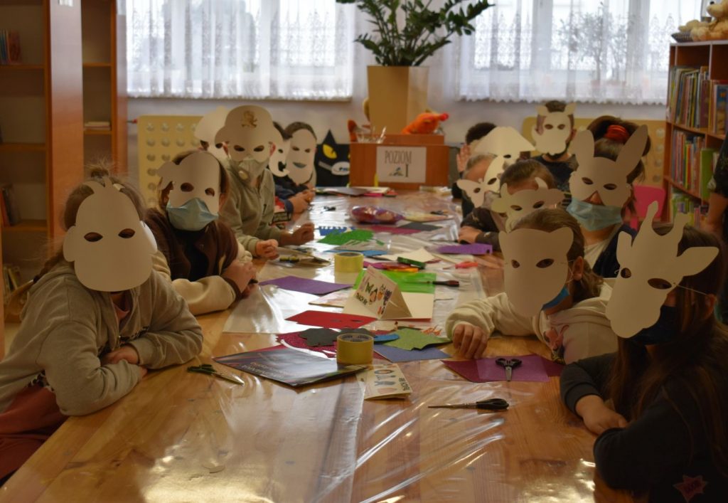 Dzieci w białych maskach siedzą przy stole, w tle dwa okna i regały na których stoją książki
