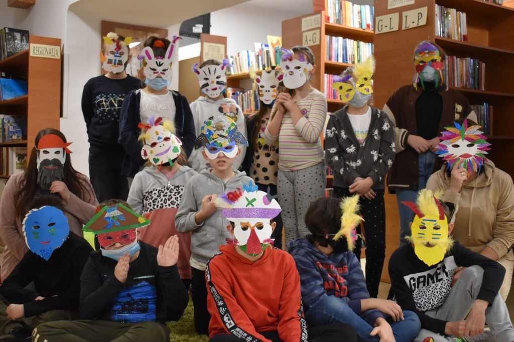Gromadka dzieci w kolorowych maskach zrobionych przez siebie. W tle regały pełne książek