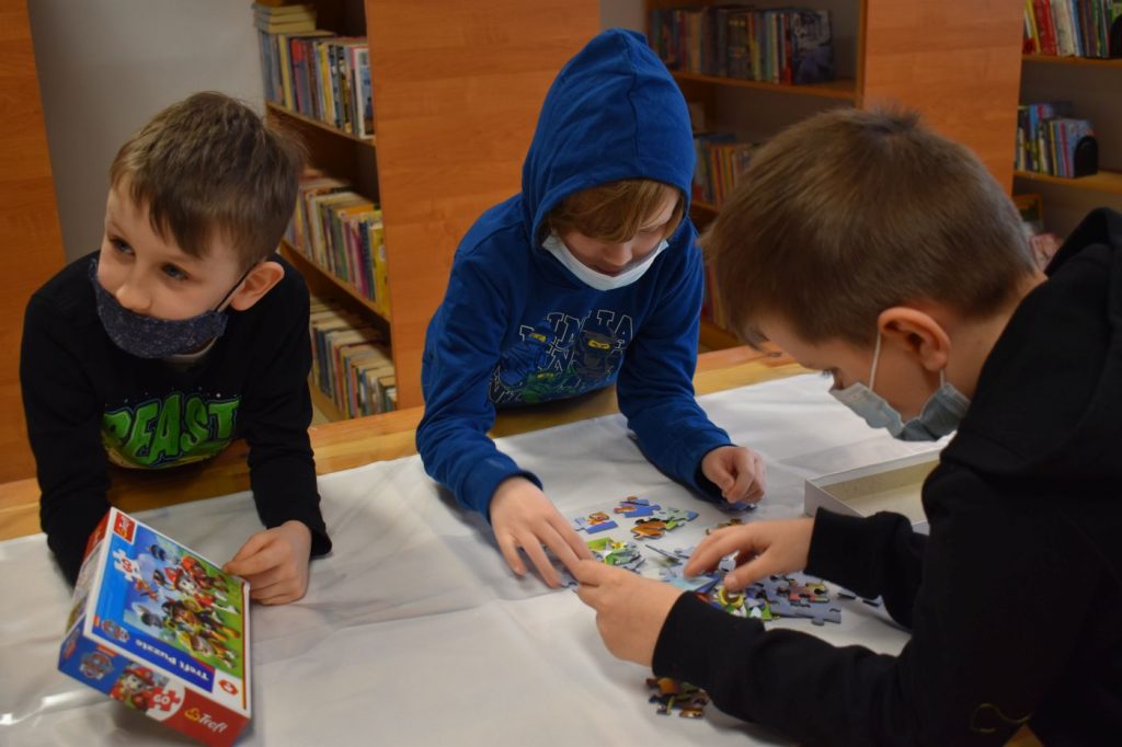 Dwóch chłopców układa puzzle, trzeci trzyma pudełko, w tle regały z książkami