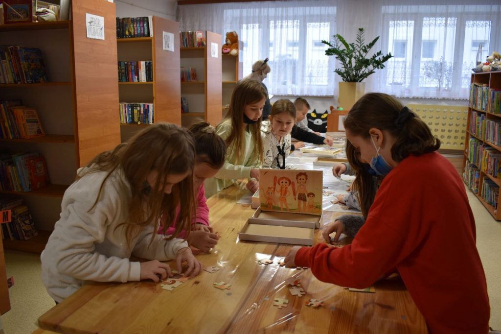 Przy stole siedzą dzieci i układają puzzle, w tle regały z książkami