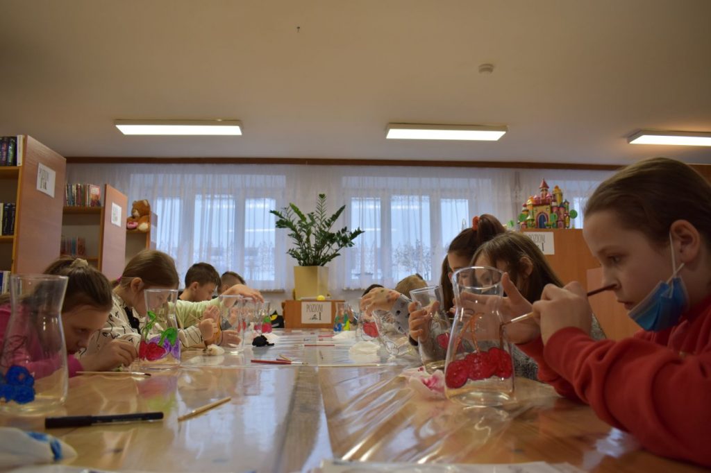 Grupa dzieci maluje szklane wazony na których widać wiśnie. W tle duży zielony kwiatek w żółtej doniczce i regały