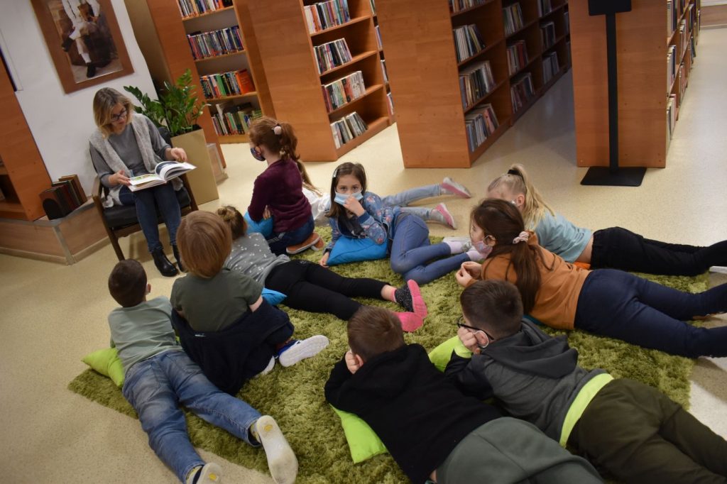 Na zielonym dywanie leżą chłopcy i dziewczęta słuchają kobiety czytającej książkę w tle regały z książkami