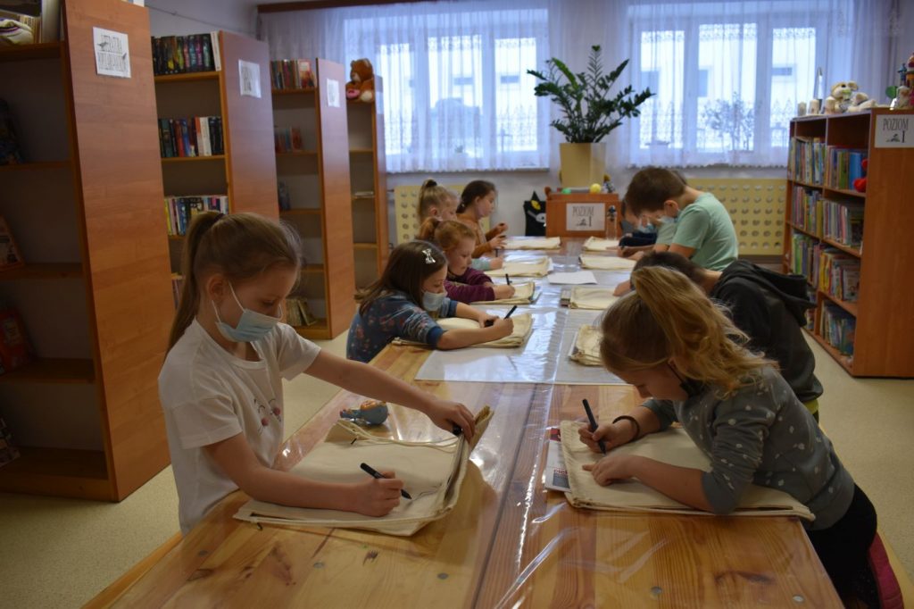 W bibliotece przy długim stole dzieci rysują na worko-plecakach w tle regały biblioteczne z książkami