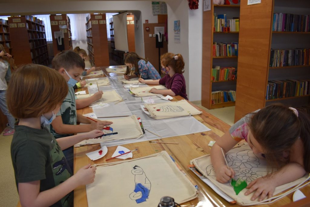 grupka dzieci maluje na materiale przy długim stole w tle regały z książkami
