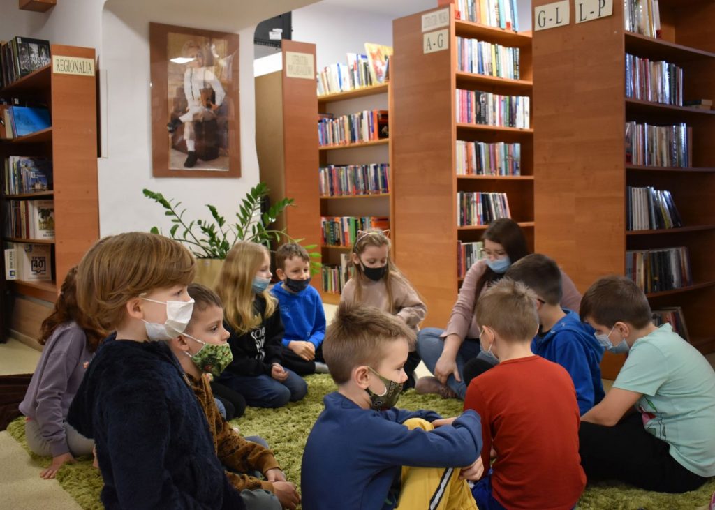 Dzieci siedzą w kółku na zielonym dywanie w tle regały z książkami