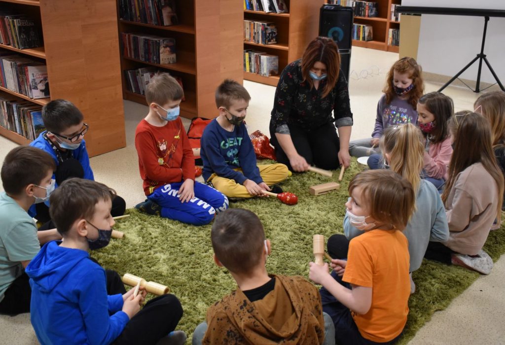 Dzieci siedzą na dywanie w ręku trzymają drewniane instrumenty za nimi regały biblioteczne z książkami