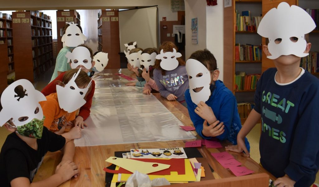 Grupka dzieci w białych maskach siedzi w bibliotece przy długim stole za nimi książki stojące na półkach