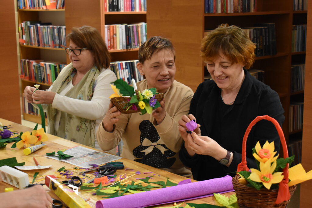 Trzy kobiety siedzą przy stole robią kwiaty z krepy, w tle regały z książkami