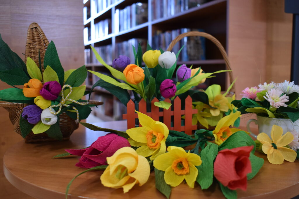 Na stoliku leżą kolorowe kwiaty z krepy. W tle półki z książkami