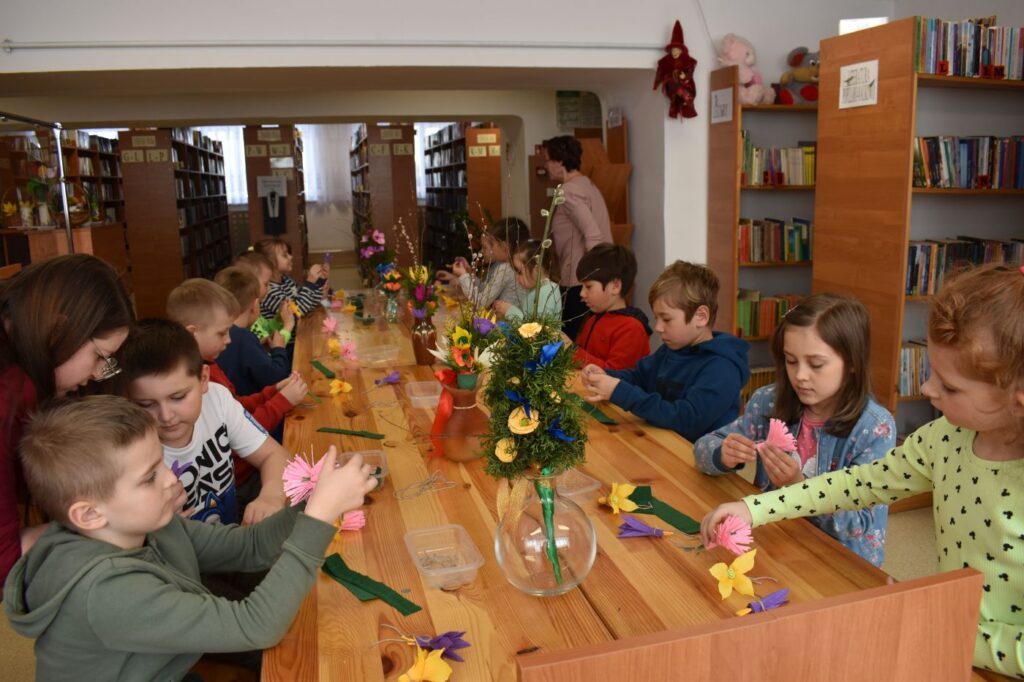 Dzieci robią kolorowe kwiaty z krepy siedząc przy stole na którym stoją kolorowe palmy. W tle regały z książkami