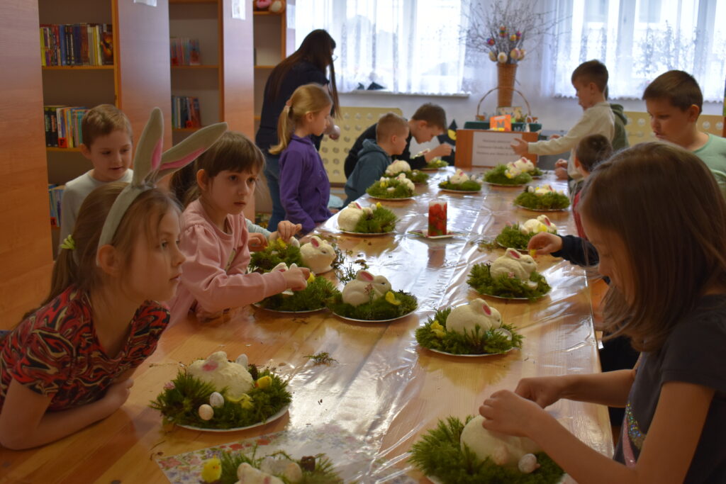 Grupa dzieci siedzi przy stole na którym stoją talerzyki na których jest zielony mech i zajączki zrobione przez dzieci. W tle regały z książkami.
