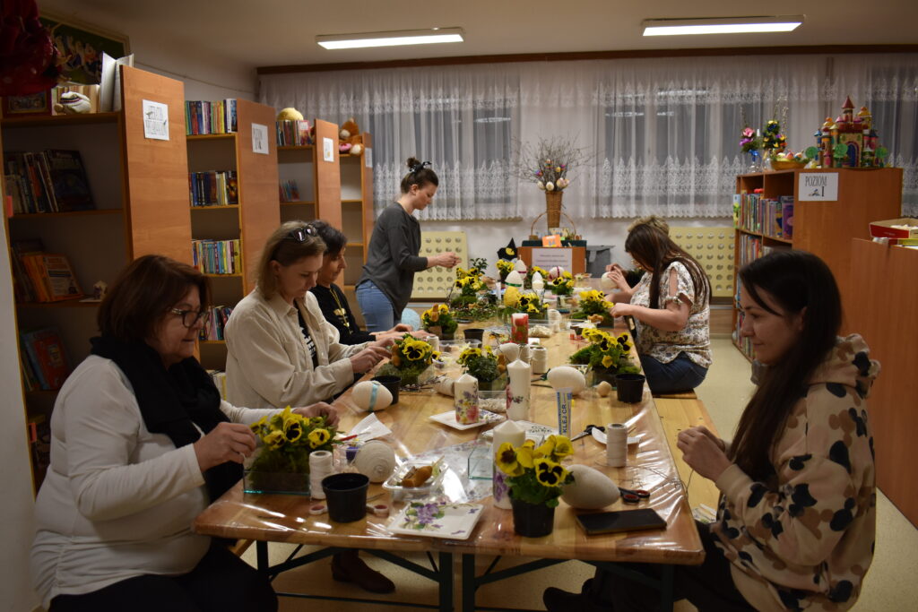 Przy drewnianym stole siedzą kobiety które robią dekoracje świąteczne z żywymi kwiatami w kolorze żółtym obol każdej leżą styropianowe jajka. W tle regały biblioteczne z książkami.